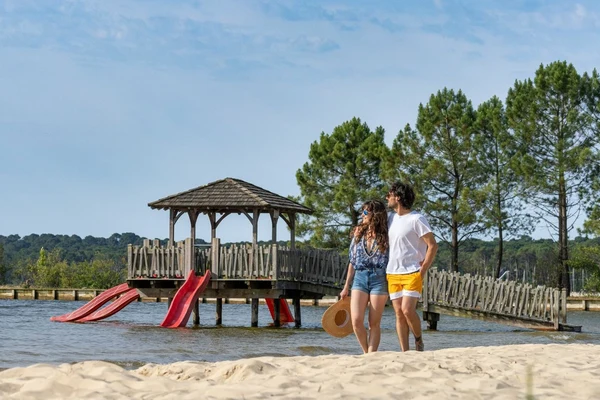 A couple strolls on a sandy lake beach near a wooden pavilion with slides; trees surround the area at Maguide