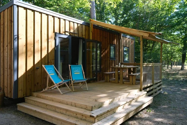 Wooden cabin has blue deck chairs on a sunlit porch amidst tall green trees at Seasonova Du Chêne