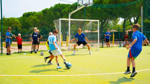Children playing soccer, kicking a ball towards the goal, watched by others, on an outdoor field surrounded by trees at Bella Italia