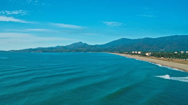 Waves gently roll toward a sandy beach, bordered by a mountainous landscape under a clear blue sky at Les Albères