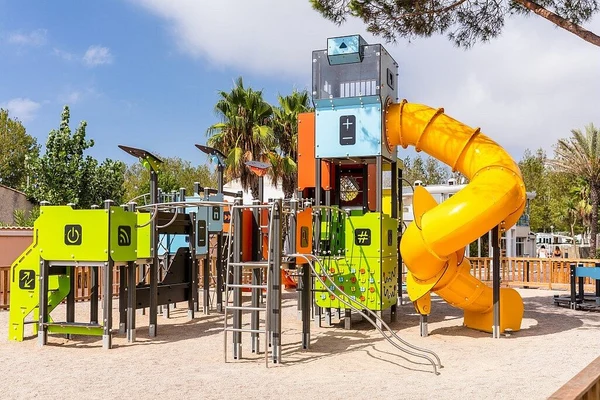 Colorful playground structure featuring slides and climbing elements on sandy ground surrounded by palm trees and buildings at Marvilla Parks Club Le Napoléon