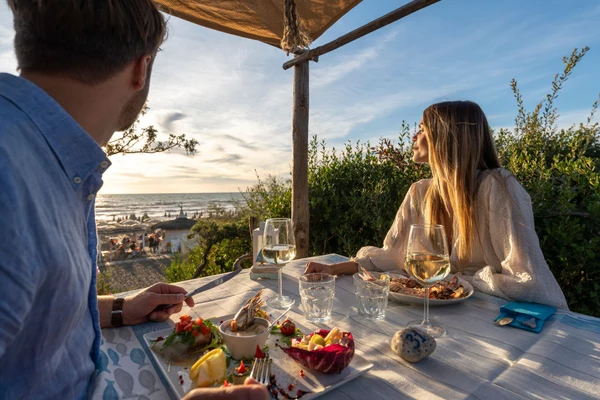 Two people enjoy a seaside dinner on a wooden table, overlooking a beach at sunset at Etruria Camping Village
