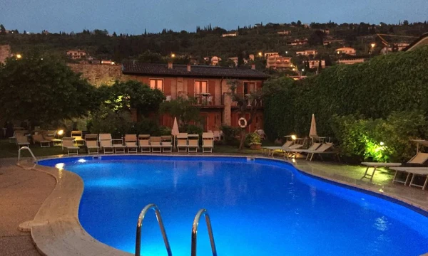 Illuminated swimming pool reflecting blue light, surrounded by lounge chairs and greenery; quaint building and hillside houses in background at Hotel Romeo