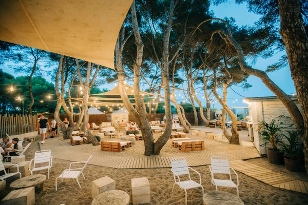 Wooden seating and tables under string lights and canvas shades amidst trees, surrounded by sand pathways and cabins at Tamarit Beach Resort