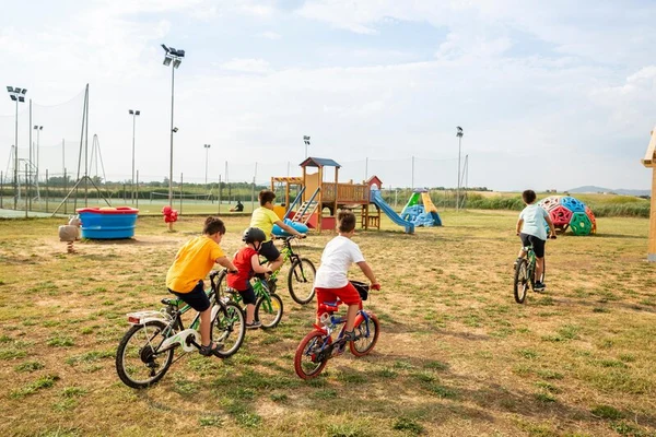 Children riding bicycles on grass near a playground with slides and climbing structures at Club Degli Amici