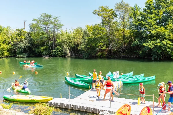 Kayakers paddle on a river, while people prepare kayaks on a dock, surrounded by lush trees at Marvilla Parks Club Le Napoléon