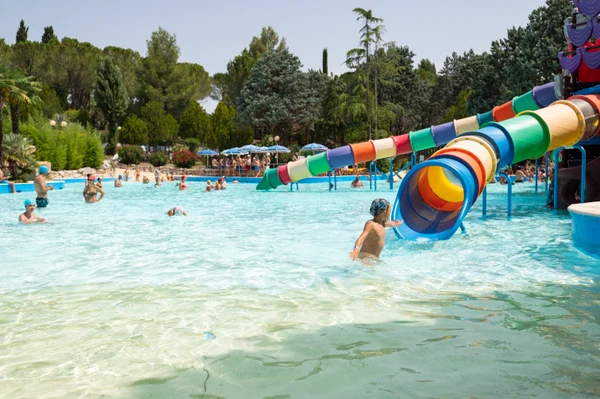 Children playing in a water park pool with a colorful slide, surrounded by trees and people lounging at Centro Vacanze Verde Azzurro