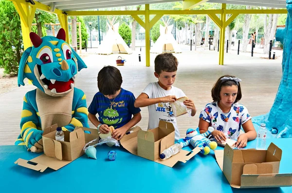 Mascot and children prepare food at a blue table under a pavilion with tents nearby at Tamarit Beach Resort