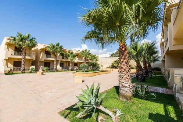 Palm trees stand tall in a sunny courtyard surrounded by beige buildings, featuring well-kept lawns and pathways at Triscina Mare
