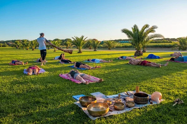 People lie on grass mats while a person plays a didgeridoo, surrounded by palm trees at Club Degli Amici
