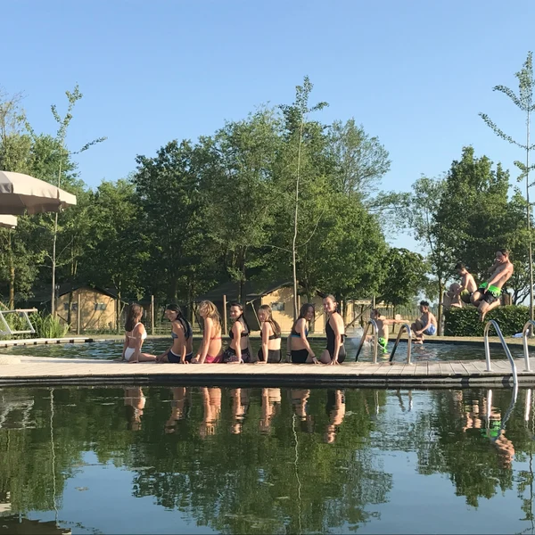 People sitting on a wooden dock, wearing swimsuits, at a natural pool with trees and cabins in the background at Agriturismo Tenuta Regina