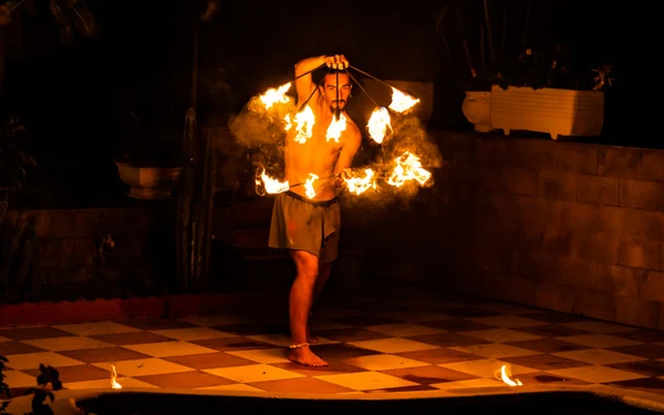 A shirtless man twirling fire poi on a tiled patio at night, creating bright flames at Triscina Mare