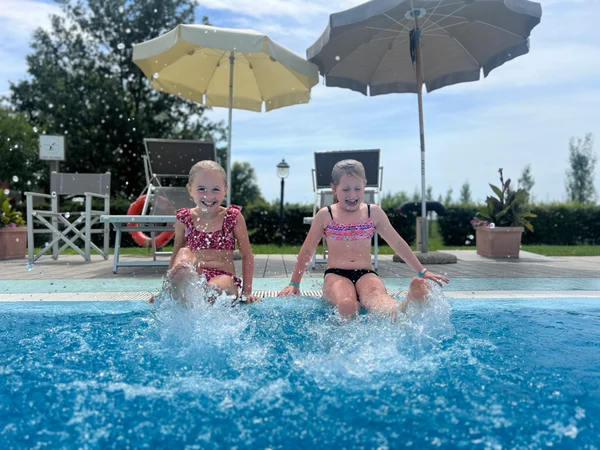 Two children splash water with their feet while sitting poolside, beneath umbrellas and surrounded by greenery at Agriturismo Tenuta Regina