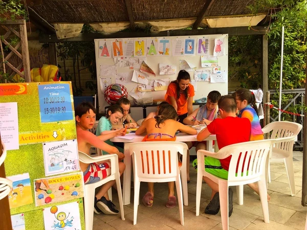 Children drawing at a table with an instructor, surrounded by artwork and informational signs at Village Torre Pendente