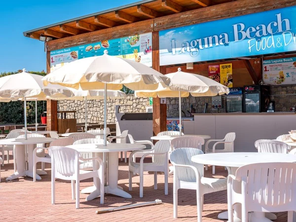 Restaurant seating area with white plastic tables and chairs under umbrellas, in front of a snack bar at Residence Eden