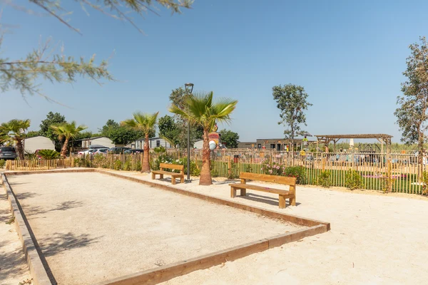 Two wooden benches facing a sandy bocce court, surrounded by palm trees, near mobile homes at Marvilla Parks Le Castellas