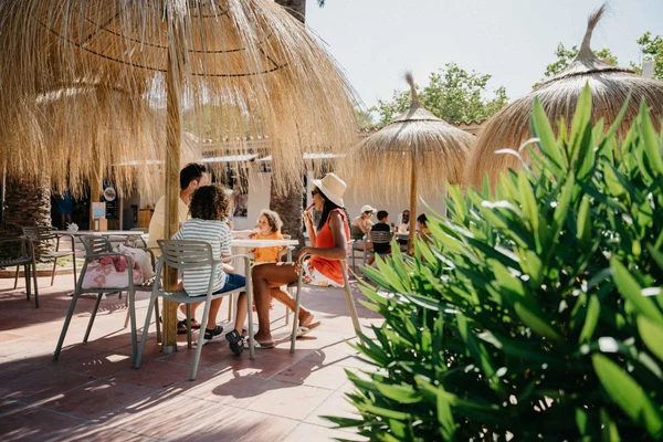 People sitting and chatting at a table under straw umbrellas, surrounded by lush greenery at Tamarit Beach Resort