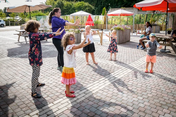 Children following an outdoor dance lesson led by an adult on a paved area with benches and umbrellas at 't Geuldal
