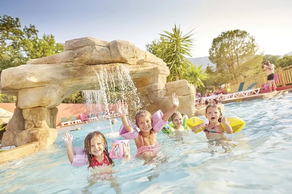 Children playing under a rock waterfall in a pool, surrounded by lounge chairs and trees at Les Albères