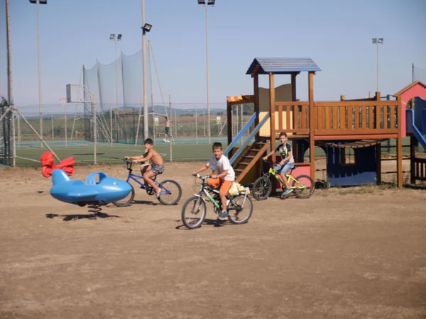 Children riding bicycles near a playground with slides and swings, adjacent to a fenced sports court at Club Degli Amici