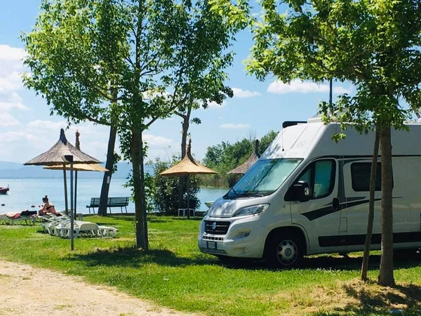 Campervan parked beside a lake, surrounded by trees and straw umbrellas; people relax by the shore at Punta Navaccia