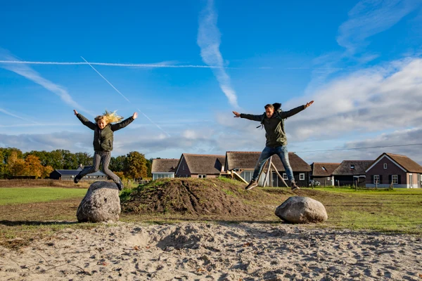 Two people jumping from rocks, arms outstretched, in a grassy area with houses and blue sky in background at De Heihorsten