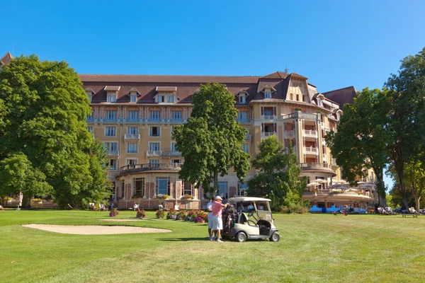 Golf cart parked on green lawn near large historic building with trees, bushes, and people in background at Seasonova Vittel