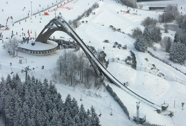 A ski jump ramp overlooks a snowy slope with skiers and snow-covered trees at Bergresort Winterberg