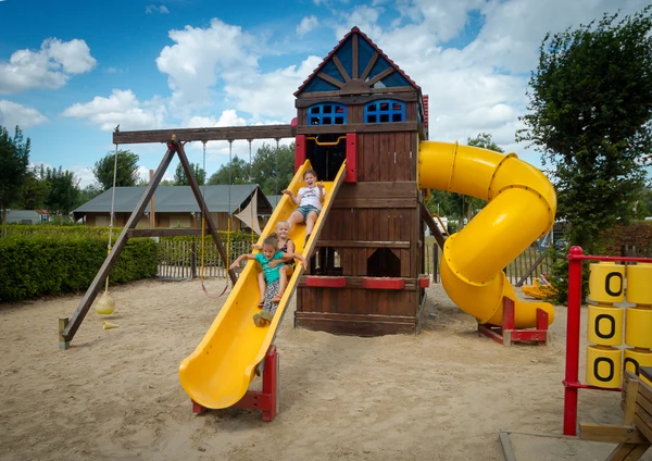 Three children slide down a yellow playground slide attached to a wooden play structure in a sandy campsite area at 't Geuldal