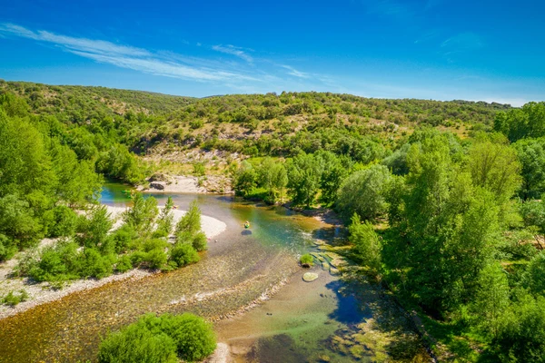 Kayakers paddling on a clear river winding through a lush, hilly forest under a bright blue sky at du Domaine d'Anglas