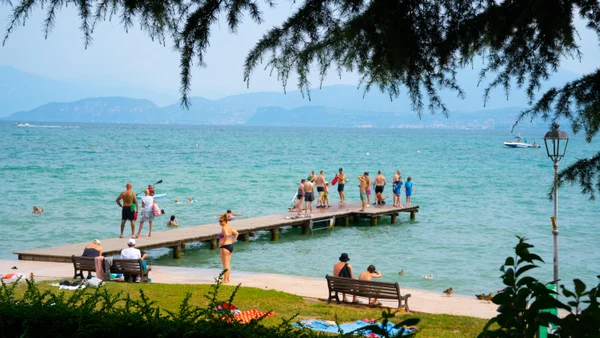 People relax and swim on a lakeside pier, surrounded by trees and distant mountains at Bella Italia