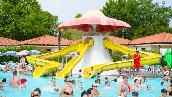 Mushroom-shaped water slide structure with children playing in a busy pool surrounded by trees and umbrellas at Bella Italia
