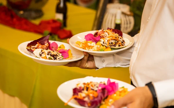 Server carrying three colorful salad plates, garnished with flowers, at a festive indoor dining area at Triscina Mare