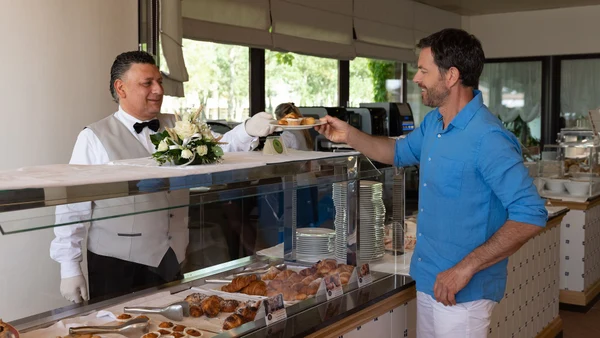 Server in a white vest hands over pastries through a glass counter to a smiling customer at Savoy Beach Hotel & Thermal Spa