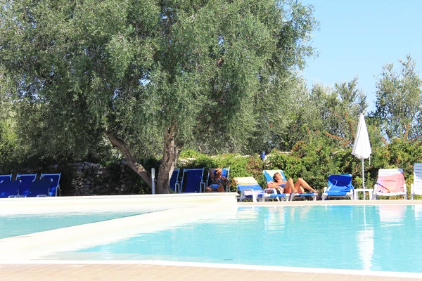 People relaxing on lounge chairs beside a pool, surrounded by trees and greenery at Porto Cesareo Camping