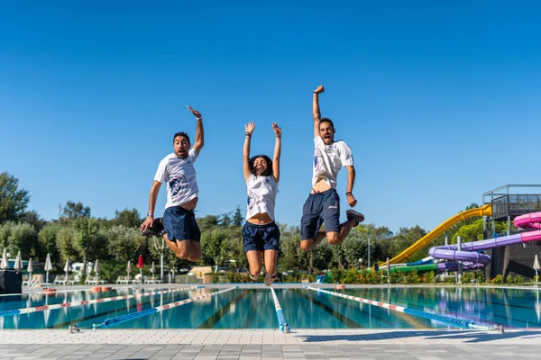 Three people in white shirts jump joyously by a pool, surrounded by green trees and colorful water slides at Marina Julia Family Camping Village