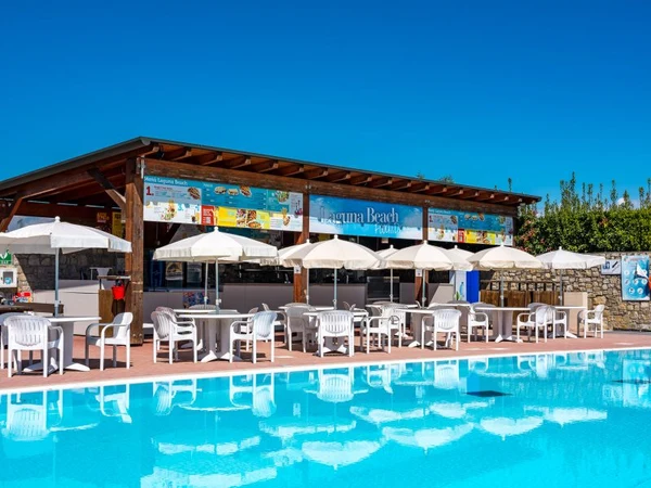 Outdoor cafe with white umbrellas and plastic chairs in front of a swimming pool under clear blue skies at Residence Eden