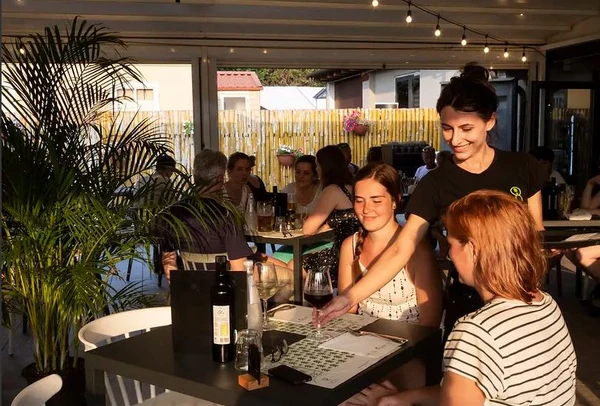 Waitress serving drinks to two smiling women at an outdoor restaurant with string lights, surrounded by plants and other patrons at hu Venezia Camping in Town