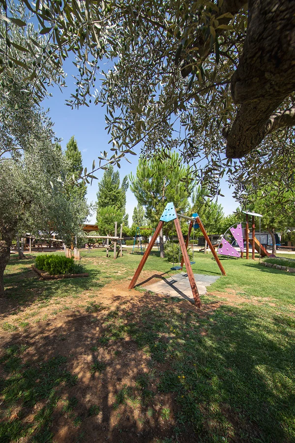 A children's swing stands in a playground surrounded by trees under a clear sky at Els Ports