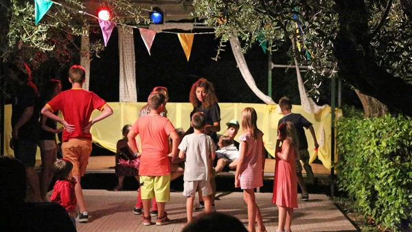 Children interacting with an adult under colorful bunting flags on a stage, surrounded by trees at Villenpark Sanghen
