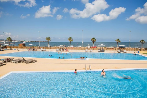 People swimming in two outdoor pools with lounge chairs nearby, overlooking the ocean and a partly cloudy sky at Taïga Delta De L'Ebre