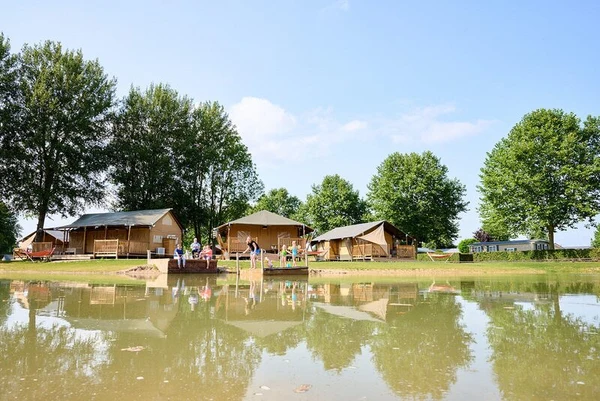 Tents stand in a grassy area beside a pond, where people gather and children play, surrounded by trees at Betuwe