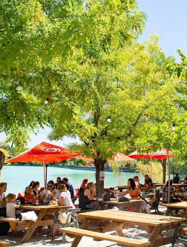 People sitting at picnic tables, eating and talking under trees with red umbrellas beside a lake at de la Bonde