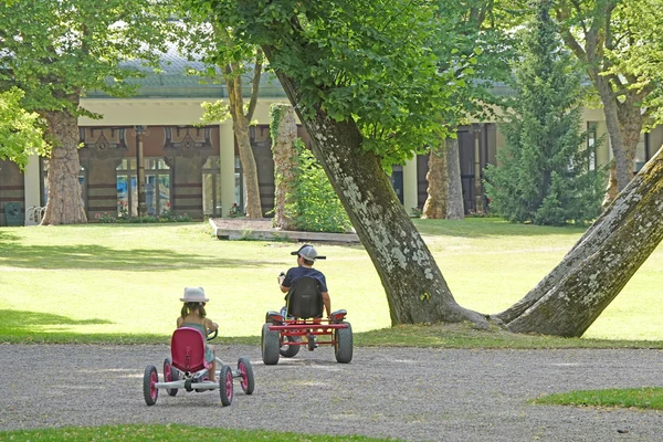 Children ride go-karts on a gravel path in a leafy park with trees and a building in the background at Seasonova Vittel
