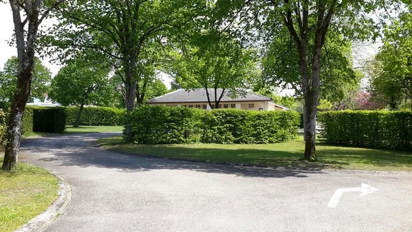 Building surrounded by lush green hedges and trees on a sunny day, with a paved road in front at Seasonova Vittel
