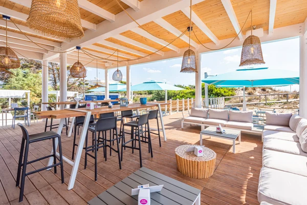 Outdoor seating area with high tables and lounge chairs on a wooden deck, surrounded by wicker lamps and umbrellas at Porto Cesareo Camping