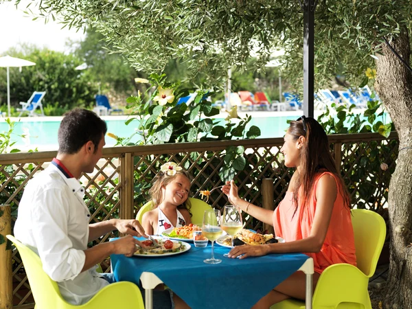 Family dining at a table, enjoying a meal with a pool and garden in the background at Porto Cesareo Camping