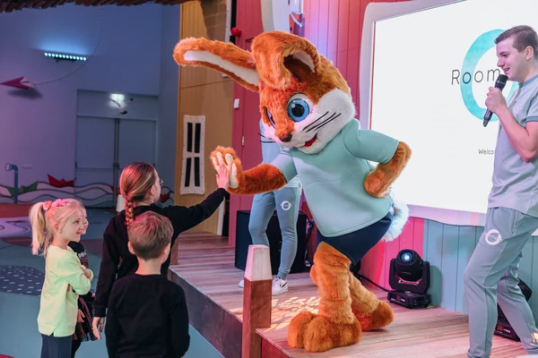 A person in a rabbit costume high-fives children on a stage, with a man holding a microphone beside them at Hunzedal
