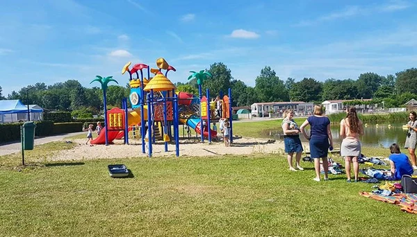 Children's playground with colorful equipment and slides, surrounded by people relaxing on grass near a pond at Betuwe