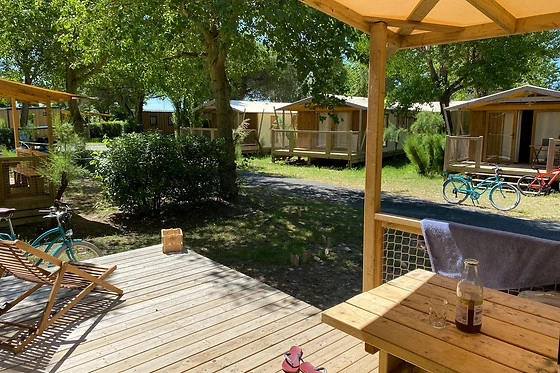 Wooden deck with chairs and table, shaded by a roof, overlooking nearby cabins and bicycles under trees at Seasonova Ile de Ré